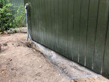 Billedet viser hjørnet af et skur i en have. Mellem skurets bund og jorden er der udlagt et jernnet, der skal bruges til rottesikring.